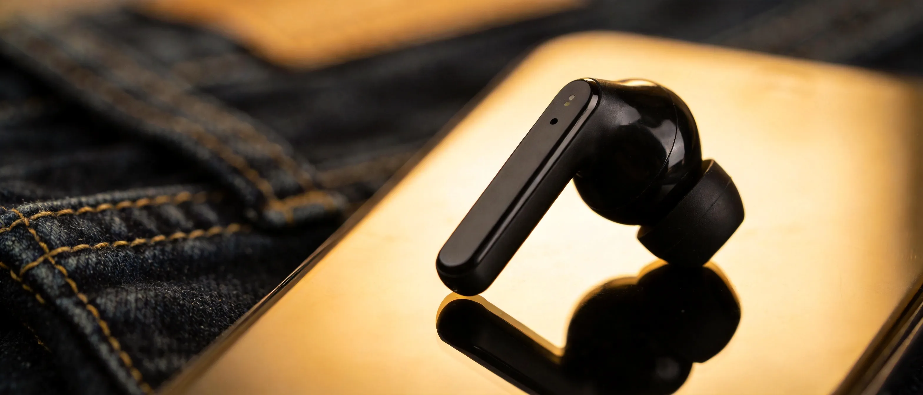 Extreme close-up macro shot of a black wireless earbud resting on a dark denim fabric, with one soft golden highlight on the glossy surface, depth of field blurring the background, futuristic and sleek mood, product advertising style.
