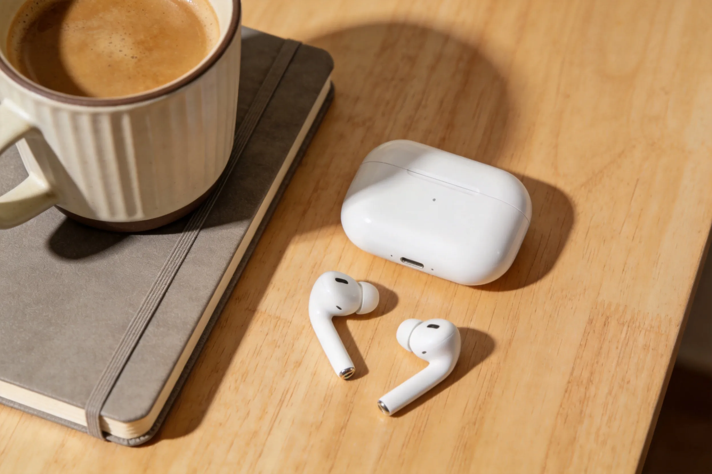 White wireless earbuds with charging case on a light wood desk, beside a coffee cup and notebook, warm indoor lighting, cozy and clean aesthetic, top-down view, 4K.