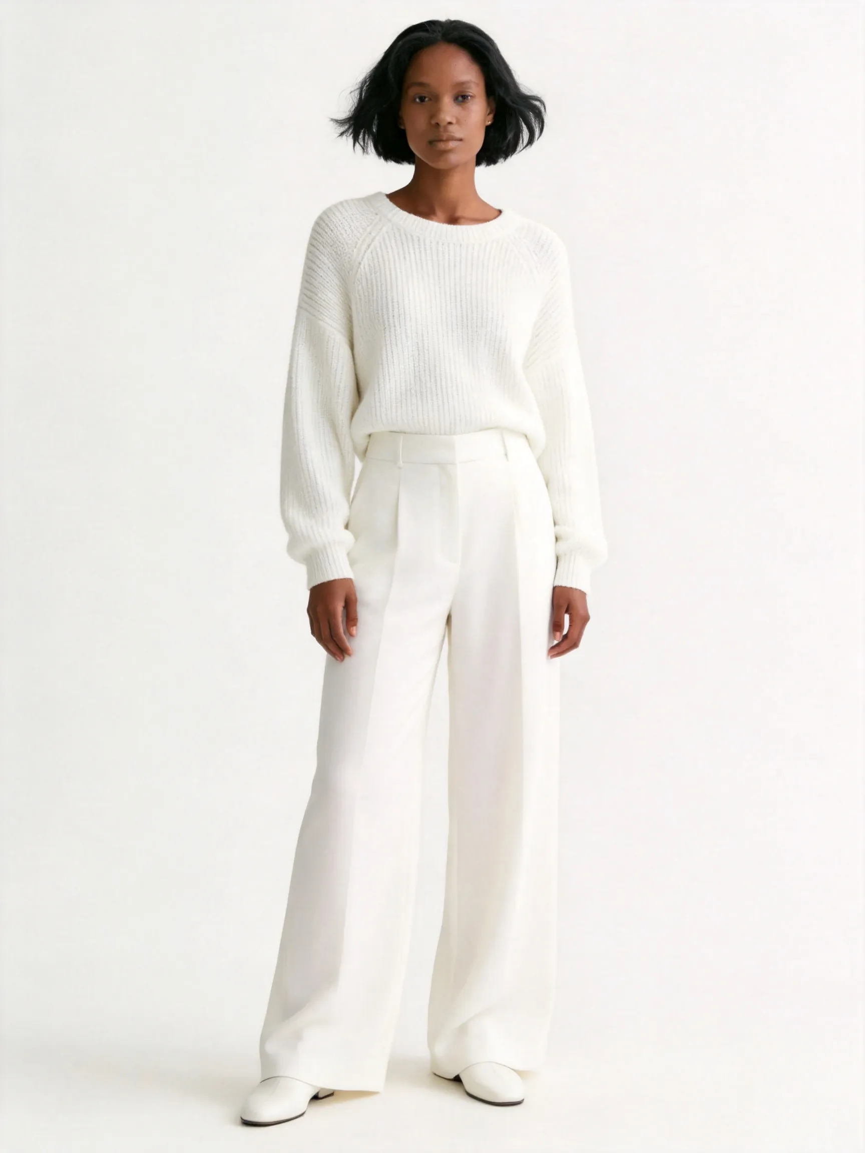 Full body shot of the woman from the reference image, standing in a natural, confident and elegant full-length posture. She features her distinctive textured black mid-part bob haircut and calm facial features. She is wearing a sophisticated all-white monochromatic ensemble: a premium chunky white knit crewneck sweater, beautifully paired with matching high-waisted fluid white wide-leg trousers that drape elegantly, and minimal white leather shoes, maintaining a cozy, luxurious, and clean aesthetic. The background is a plain, seamless studio backdrop in a soft off-white/cream hue with gentle diffused lighting, keeping the environment and muted color tone completely consistent with the reference image. High-end fashion editorial photography style, sharp focus, 8k resolution, photorealistic --ar 3:4 --v 6.0