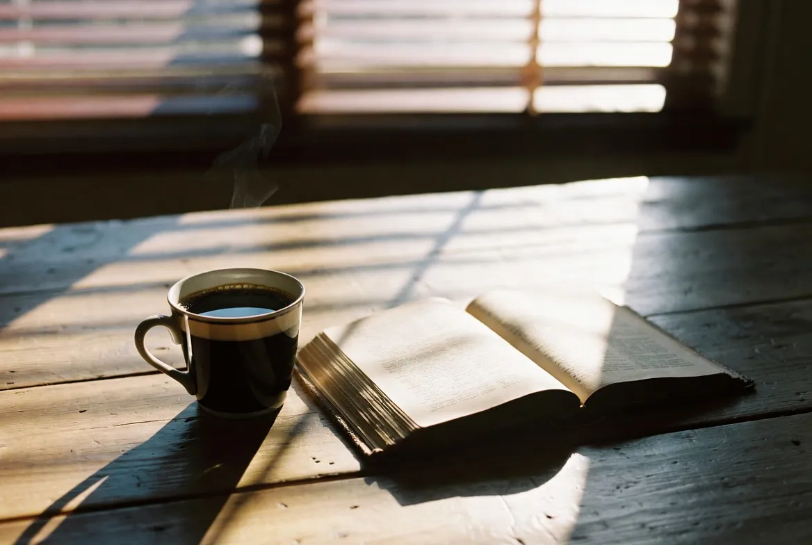 Minimalist still life photography, a cup of black coffee with steam rising next to an open vintage book on a rustic wooden table, warm morning sunlight casting striped shadows through venetian blinds, shallow depth of field focusing on the coffee cup, film grain, moody and cozy atmosphere, Kodak Portra 400 style, 4k, hyper-realistic texture.