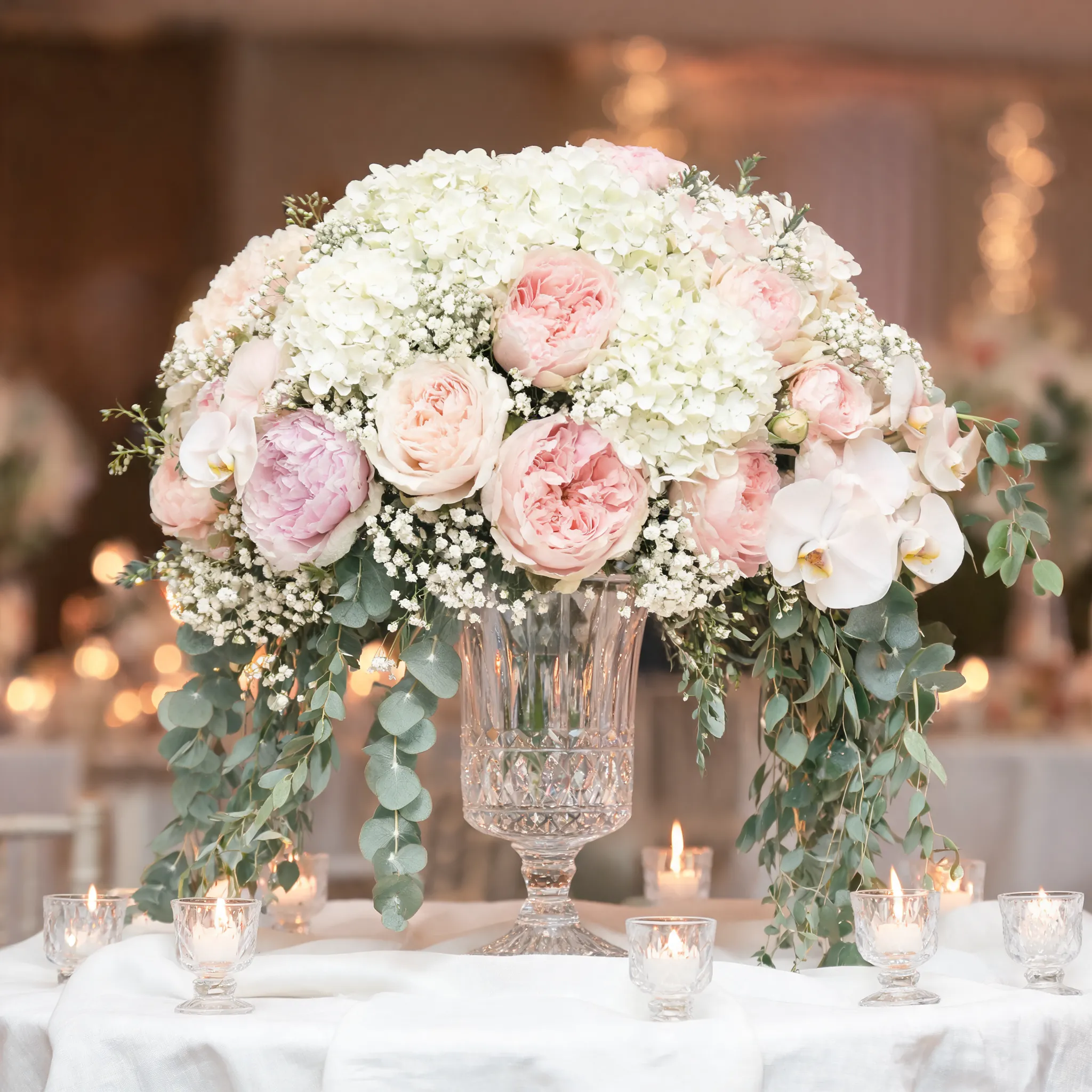 A luxurious wedding floral centerpiece, overflowing with white hydrangeas, blush roses, peonies, garden roses, orchids, baby's breath, eucalyptus, and cascading greenery, arranged in a crystal vase, elegant symmetrical composition, romantic atmosphere, luxury wedding florist design, soft candlelight, premium floral styling, realistic flower textures, magazine-quality photography, photorealistic, 8K