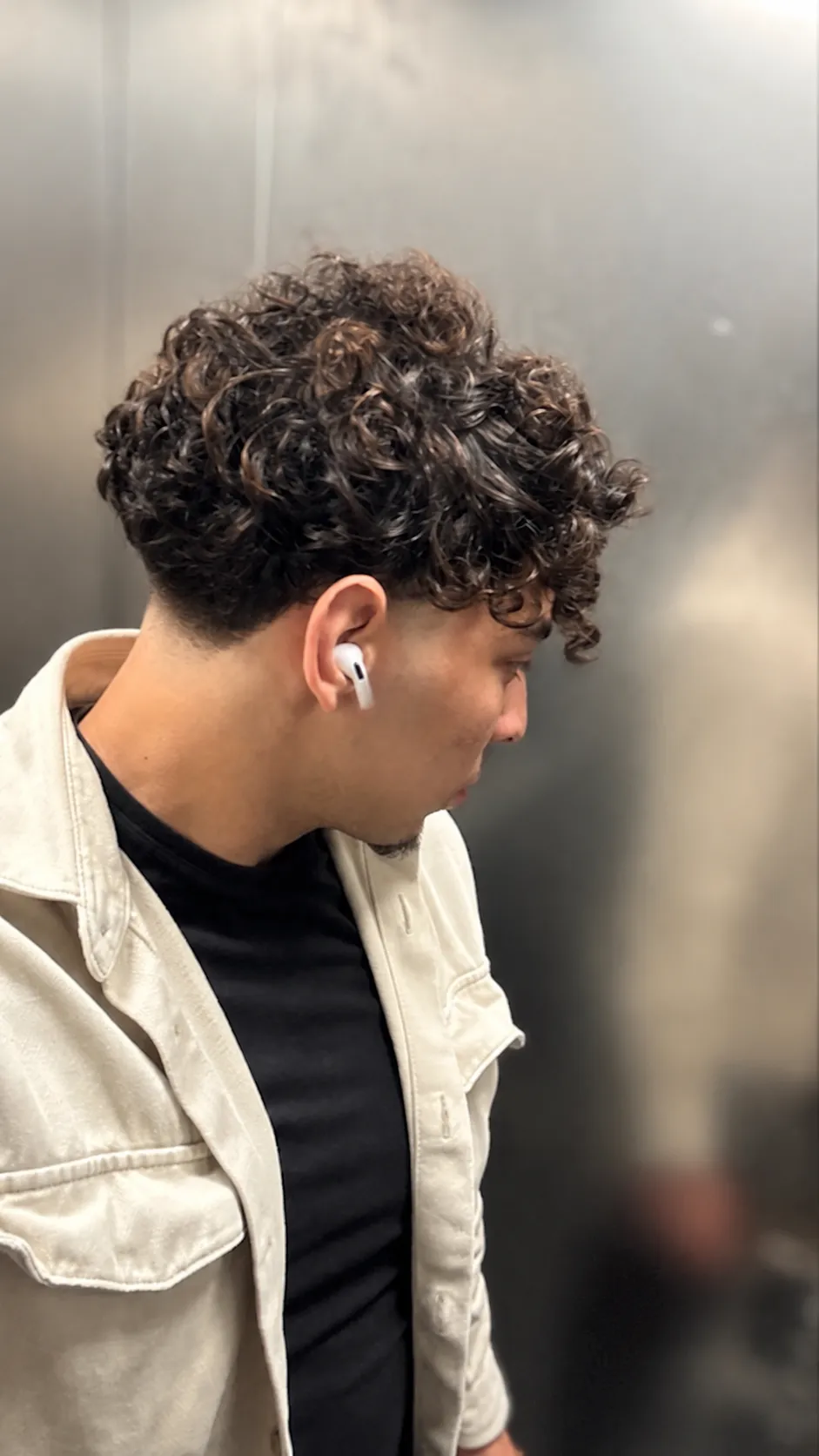 Subject & Style: A young man with dark, curly hair wearing a light jacket over a black t-shirt. Hair Color & Highlights Specification: • Proportions: 70% natural black hair (dominant base color) and 30% highlighted strands. • Shade / Tone: Warm chocolate chestnut brown that contrasts subtly with the black base-visible and luminous, but not too light or blonde. • Technique & Distribution: • Seamless balayage technique with a soft, gradual blend and no harsh lines or visible roots. • Highlights start mid-shaft along the length of the curls. • Even distribution across the entire head (top, sides, and back-not just the front strands). • Overall Effect: Adds depth, dimension, and definition to the curly texture while maintaining a dark, natural, and sophisticated look.