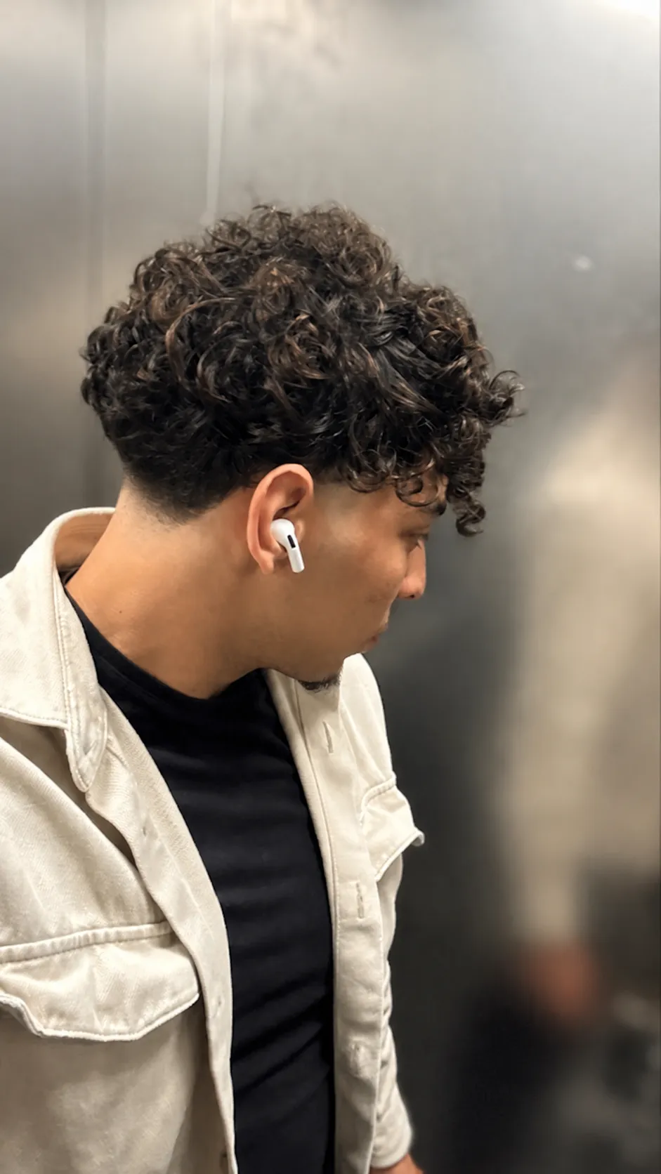 Subject & Style: A young man with dark, curly hair wearing a light jacket over a black t-shirt. Hair Color & Highlights Specification: • Proportions: 70% natural black hair (dominant base color) and 30% highlighted strands. • Shade / Tone: Deep chestnut brown (châtain foncé). The highlights should be a deep, rich brown that is clearly distinguishable from the natural black base, providing a distinct contrast without being light brown or honey/ caramelized. • Technique & Distribution: • Seamless balayage technique with a soft, gradual blend and no harsh lines or visible roots. • Highlights start mid-shaft along the length of the curls. • Even distribution across the entire head (top, sides, and back—not just the front strands). • Overall Effect: Adds depth, dimension, and definition to the curly texture while maintaining a dark, natural, and sophisticated look.