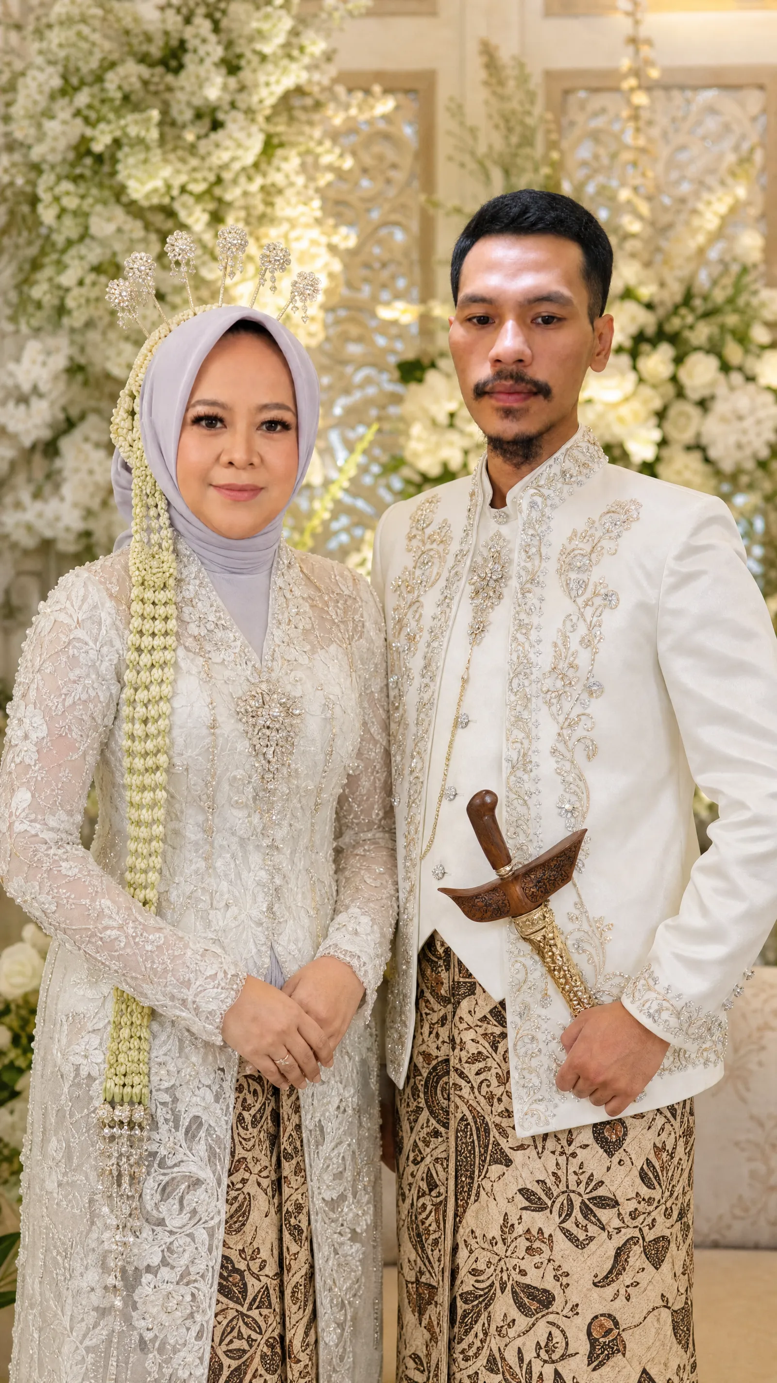 PROMPT UTAMA — PENGANTIN JAWA PUTIH GOLD ULTRA REALISTIS preservation / face fidelity sangat tinggi” sebagai prioritas tertinggi. Gunakan foto yang diunggah sebagai referensi visual utama. Pertahankan identitas wajah asli kedua orang semaksimal mungkin. Jangan membuat wajah baru, jangan mengganti karakter wajah, jangan mengubah bentuk wajah, mata, hidung, bibir, rahang, alis, warna kulit, atau ciri khas wajah asli. Buat sebuah foto pernikahan Jawa modern yang sangat realistis dan elegan, menampilkan sepasang pengantin berdiri berdampingan menghadap kamera dalam pose formal dan natural. PRIA: mengenakan busana pengantin Jawa modern berwarna putih ivory dengan bordir dan ornamen warna silver-gold, berupa beskap/jas pengantin putih berpotongan rapi dan mewah. Detail bordir floral dan ornamen tradisional berwarna champagne gold dan silver menghiasi bagian dada, kerah, manset, serta sisi depan pakaian. Mengenakan kain batik Jawa motif klasik warna cream, cokelat muda, dan cokelat tua, dengan pola batik yang sangat detail dan realistis. Tambahkan aksesori keris Jawa tradisional berwarna cokelat kayu dengan warangka ukiran elegan yang terselip di bagian pinggang. HAPUS SONGKOK DAN BLANGKON. Jangan menggunakan songkok, peci, blangkon, atau penutup kepala modern. Gunakan aksesori kepala Jawa yang sangat sederhana dan elegan, atau rambut ditata rapi secara natural sesuai karakter wajah asli. WANITA: mengenakan kebaya pengantin Jawa putih ivory, sangat mewah tetapi tetap anggun dan feminin. Kebaya memiliki material lace/transparan yang sangat halus dengan bordir floral tiga dimensi, payet kecil, kristal dan ornamen silver-champagne gold. Siluet kebaya mengikuti bentuk tubuh secara elegan dan natural, tidak berlebihan. Bagian bawah menggunakan kain batik Jawa klasik dengan perpaduan warna cream, cokelat, dan cokelat tua. Rambut wanita ditata menjadi sanggul Jawa tradisional yang sangat rapi, dihiasi aksesori pengantin Jawa berwarna silver dan kristal. Tambahkan roncean mela