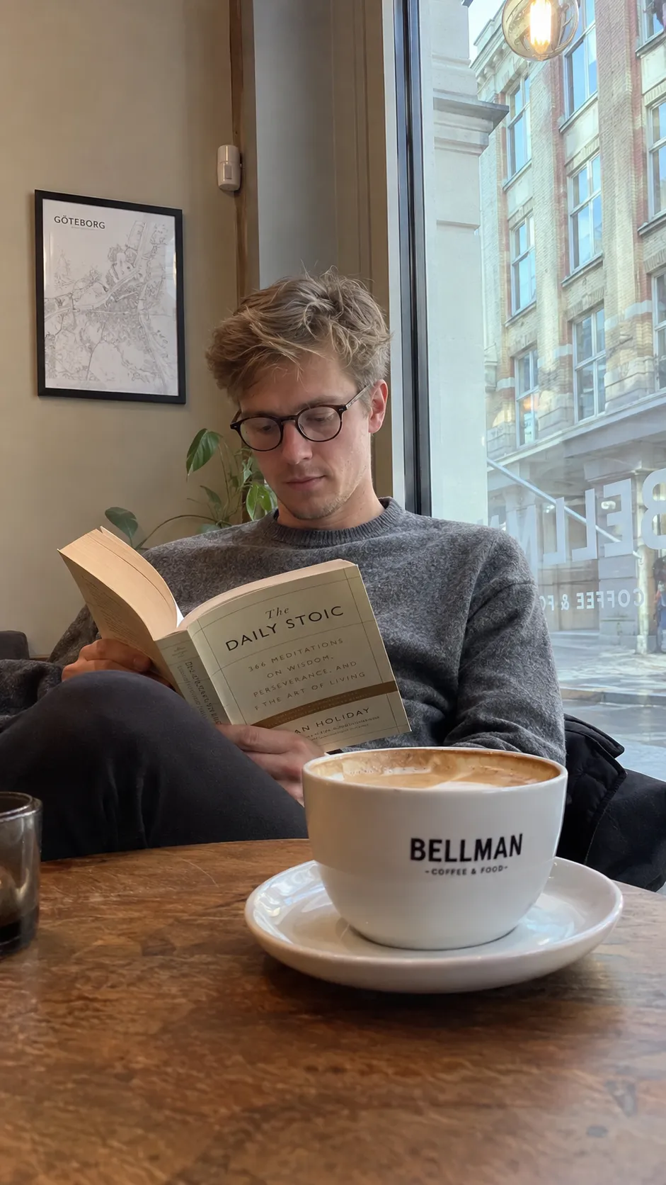 A realistic casual smartphone photo of a young Scandinavian man sitting alone in a cozy coffee shop. He wears a simple gray sweater and glasses, reading a book beside a large window. Natural morning light enters the cafe, coffee cup on the wooden table, slightly messy hair, relaxed expression, imperfect everyday composition. Captured on an iPhone camera, realistic exposure, natural skin texture, authentic lifestyle photography.