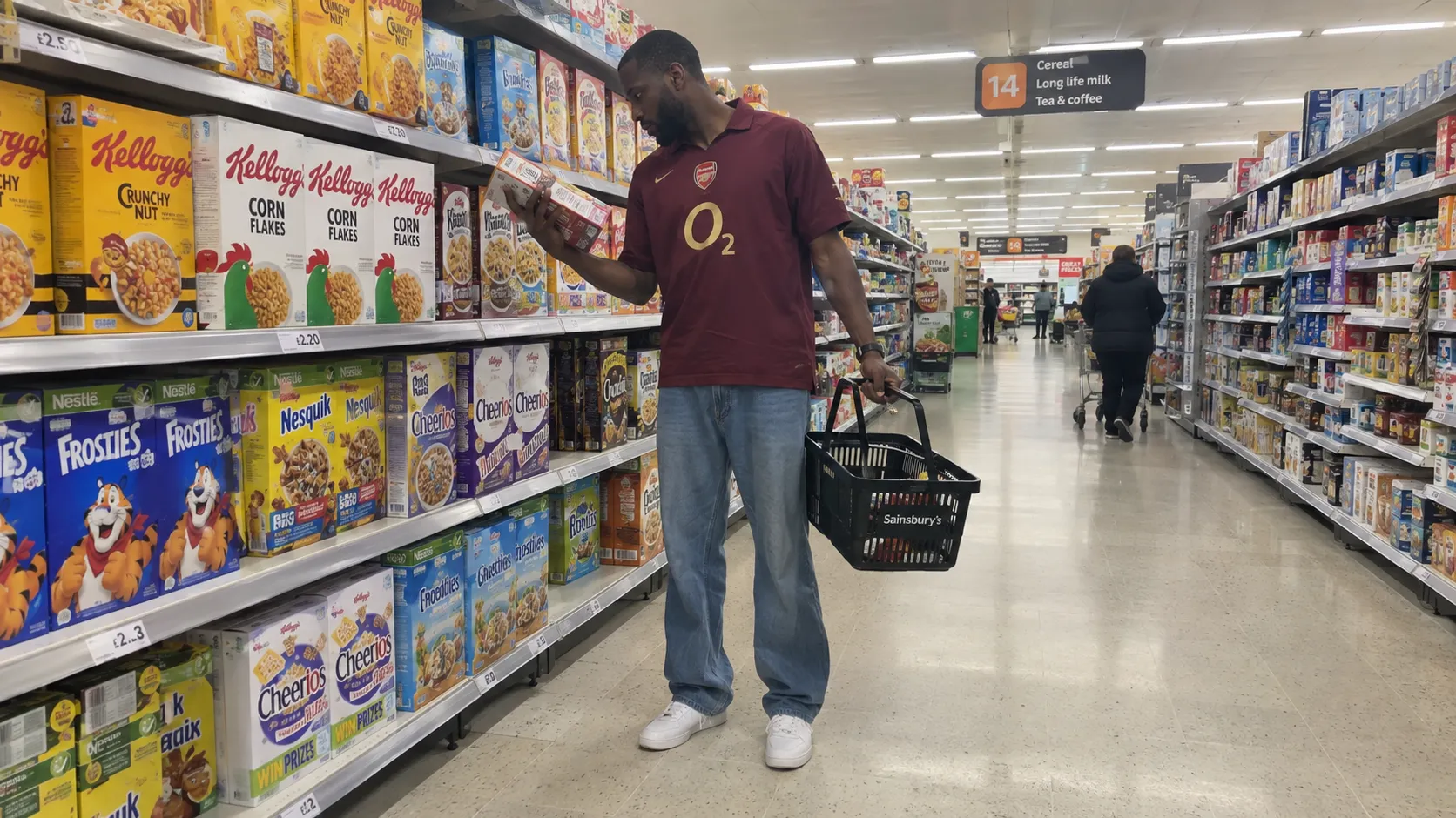 An unposed smartphone photograph of a tall Black British man standing in the cereal aisle of a modern supermarket, wearing loose jeans, a vintage football jersey and white sneakers, holding a shopping basket with one hand while reading a product label with the other, fluorescent supermarket lighting, slightly awkward natural posture, shelves extending into the background, subtle smartphone computational photography, realistic skin texture, mild lens distortion, imperfect framing, authentic everyday life rather than commercial photography.