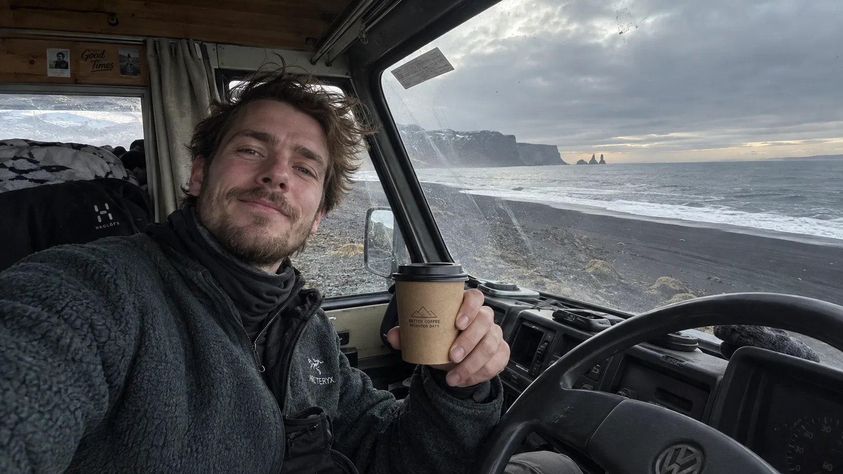 A spontaneous front-camera selfie of a young Icelandic man sitting inside an old camper van parked beside a black volcanic beach, messy windblown hair, slightly tired but happy expression, fleece jacket and hiking clothes, one hand holding a paper coffee cup, enormous dark ocean and distant cliffs visible through the windshield, overcast daylight, realistic smartphone exposure, slightly wide-angle facial distortion, fingerprints and dust visible on the windshield, authentic travel snapshot.