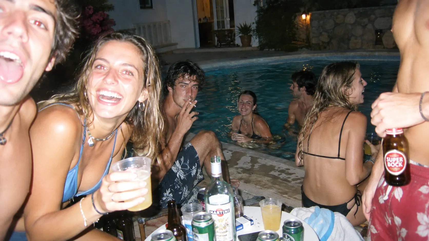 A chaotic candid photograph taken with a cheap disposable camera at a summer house party in Portugal, a group of young European friends crowded around a small outdoor swimming pool, one woman laughing directly toward the camera while another person is partially cut off by the frame, harsh built-in flash, red-eye artifacts, slight overexposure, strong shadows, cheap film grain, imperfect focus and accidental framing, authentic early-2000s party snapshot aesthetic.