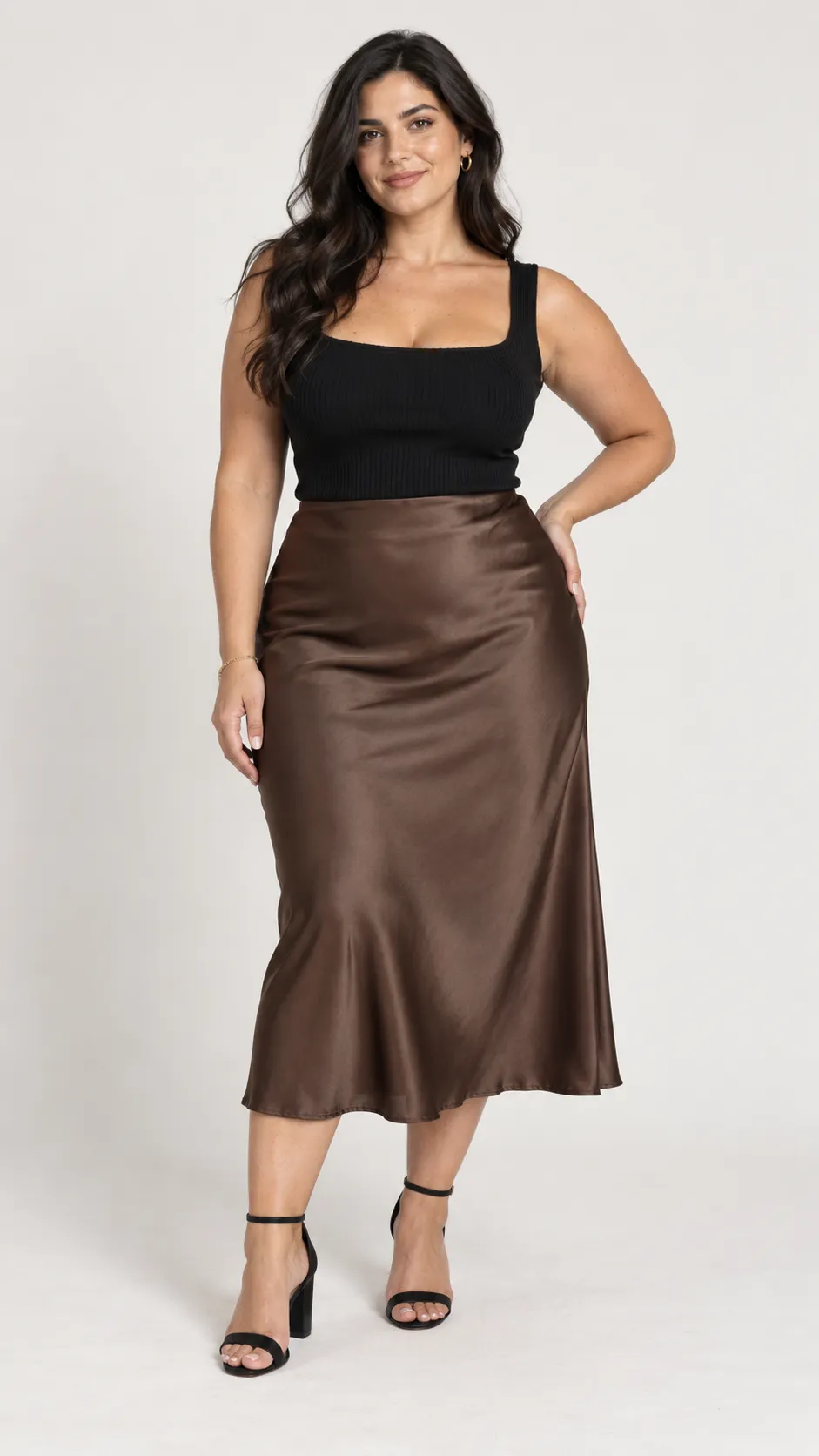 Create a photorealistic full-body fashion catalog image of a 30-year-old Mediterranean woman, approximately 168 cm tall, with a naturally curvy, plus-size figure, full hips, a defined waist, strong thighs, warm olive skin, deep brown eyes, and long dark espresso hair styled in loose glossy waves. She wears a fitted black square-neck knit top, a high-waisted chocolate-brown satin midi skirt, small gold hoop earrings, a delicate gold bracelet, and elegant black block heels. Her stance is confident but relaxed, with realistic weight distribution and natural shoulder alignment. Use sophisticated neutral makeup and authentic skin texture, including subtle pores and natural skin variations. The outfit should fit her body realistically without hiding or exaggerating her curves. Soft diffused studio lighting, clean off-white background, gentle floor shadow, detailed satin sheen and knit texture, premium fashion e-commerce photography, realistic anatomy and hands, complete head-to-toe framing, vertical 9:16 composition. No slimming distortion, no text, no watermark.