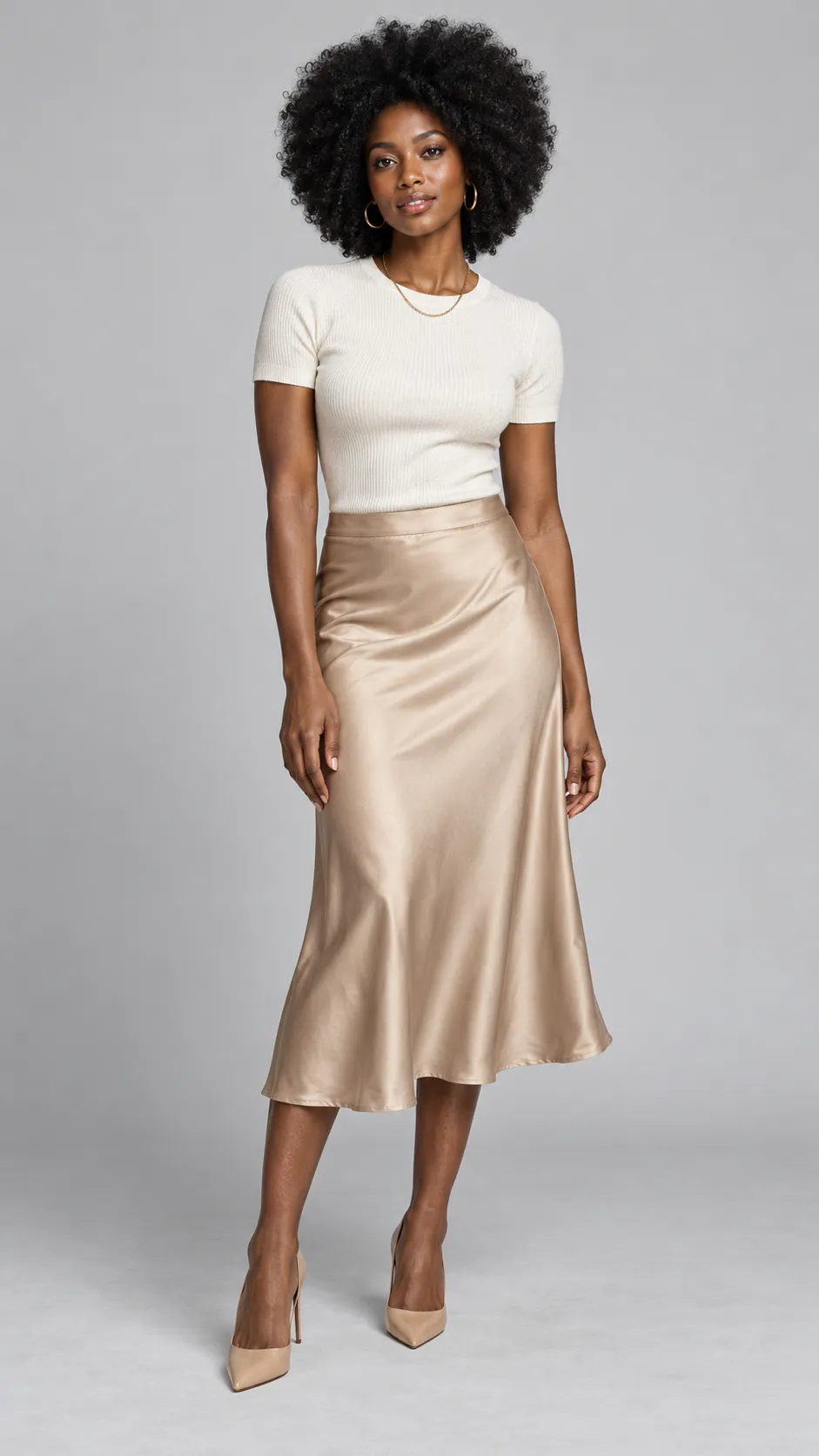 Produce a premium photorealistic full-body studio fashion portrait of a 28-year-old Black woman, approximately 175 cm tall, with a lean athletic build, deep rich brown skin, dark expressive eyes, and natural black coily hair shaped into a voluminous rounded afro. Her facial features are distinctive and elegant, with defined cheekbones and naturally full lips. She wears a fitted ivory short-sleeve knit top, a flowing warm-beige satin midi skirt, gold hoop earrings, a fine gold chain necklace, and pointed nude heels. Her pose is poised and natural, with one knee slightly relaxed and both arms resting comfortably. Preserve the richness of her skin tone without overexposure or gray color casts; retain realistic skin highlights, fine texture, and individual hair coils. Use a seamless pale-gray background, balanced softbox lighting, subtle shadows, accurate fabric reflections, luxury department-store catalog photography, realistic body proportions, full body and both shoes visible, vertical 9:16 aspect ratio. No text, no watermark, no artificial skin smoothing.