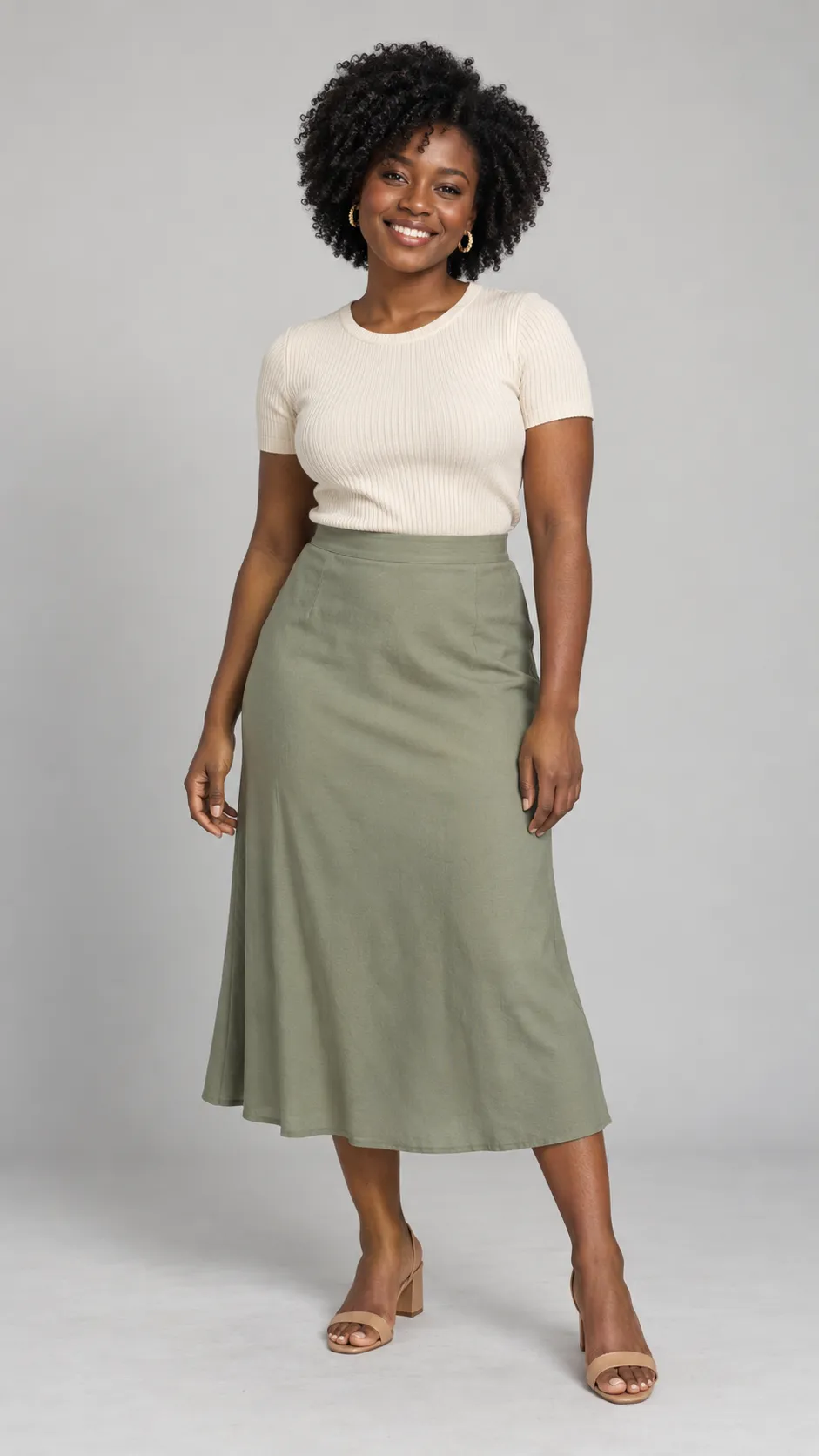 Generate a photorealistic full-body studio portrait of a 25-year-old Black woman, approximately 157 cm tall, with a petite yet naturally curvy build, deep warm-brown skin, dark eyes, and short-to-medium natural coily hair shaped into a soft rounded silhouette. She wears a cream ribbed short-sleeve top, a high-waisted muted-sage midi skirt, delicate gold earrings, and low nude block heels. Her expression is bright and genuine, with relaxed shoulders and a natural standing pose. Ensure that her skin tone remains deep and richly detailed under studio lighting, with realistic highlights and visible fine texture. The outfit must fit her petite proportions correctly, with a naturally placed waistline and realistic skirt length. Soft diffused lighting, seamless light-gray background, accurate fabric detail, subtle floor shadows, premium inclusive e-commerce fashion photography, full head-to-toe composition with shoes visible, vertical 9:16. No text, no watermark, no anatomical distortion.