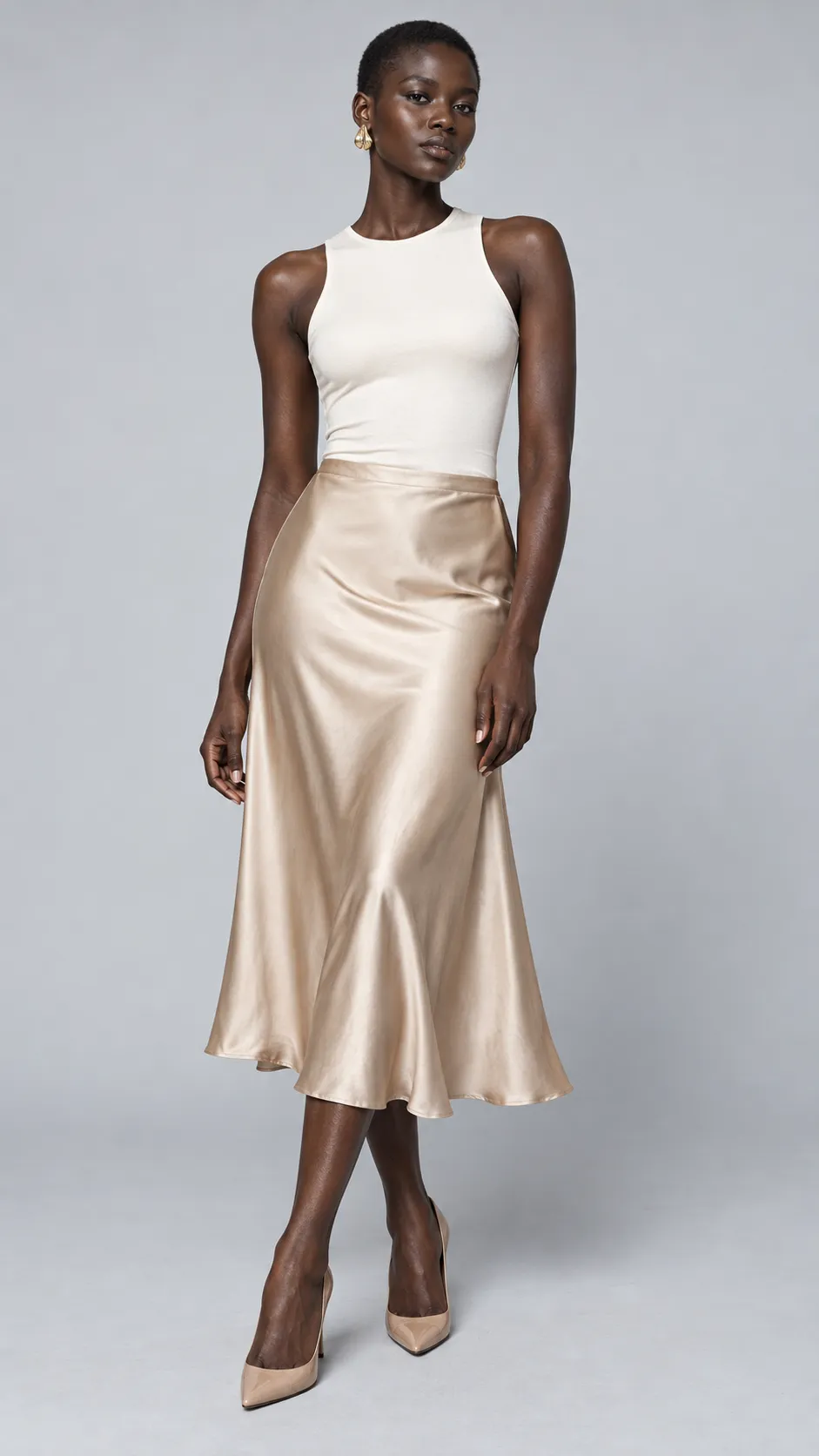 Produce a striking photorealistic full-body fashion studio image of a 28-year-old Black woman, approximately 174 cm tall, with a slim athletic silhouette, very deep ebony-brown skin, dark expressive eyes, and an ultra-short natural buzz cut. Her facial structure is distinctive, with high cheekbones and a strong jawline. She wears a minimalist ivory fitted top, a flowing champagne-beige satin midi skirt, small sculptural gold earrings, and pointed nude heels. Her expression is composed and editorial, while her body language remains natural rather than rigid. Carefully expose the dark skin so it retains rich tonal depth, subtle highlights, and realistic texture without crushed shadows. Use a seamless pale-gray background, soft balanced studio lighting, accurate satin reflections, realistic hands and limb proportions, sophisticated luxury catalog photography, complete head-to-toe framing, vertical 9:16. No text, no watermark, no artificial skin brightening.