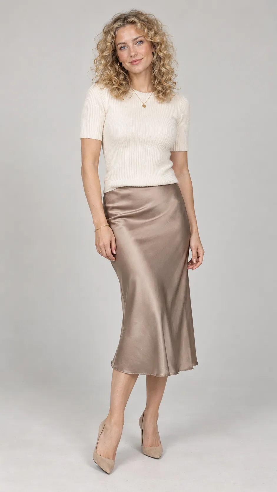 Create a high-resolution photorealistic full-body fashion portrait of a 26-year-old Dutch woman, approximately 171 cm tall, with a medium-slim figure, fair neutral skin, light gray-blue eyes, and voluminous natural golden-blonde curls falling to her shoulders. She wears a fitted cream knit top, a warm taupe satin midi skirt, delicate gold jewelry, and beige pointed heels. Her curls should have varied strand sizes, realistic volume, and a few flyaway hairs rather than looking like a uniform wig. She stands with one knee slightly bent and a gentle, confident expression. Natural makeup, visible skin texture, balanced facial features, accurate hands, and realistic body proportions. Soft commercial studio illumination, light warm-gray background, subtle floor contact shadows, precise knit and satin textures, premium contemporary fashion catalog photography, full head-to-toe view, vertical 9:16 aspect ratio. No text, no watermark, no excessive retouching.