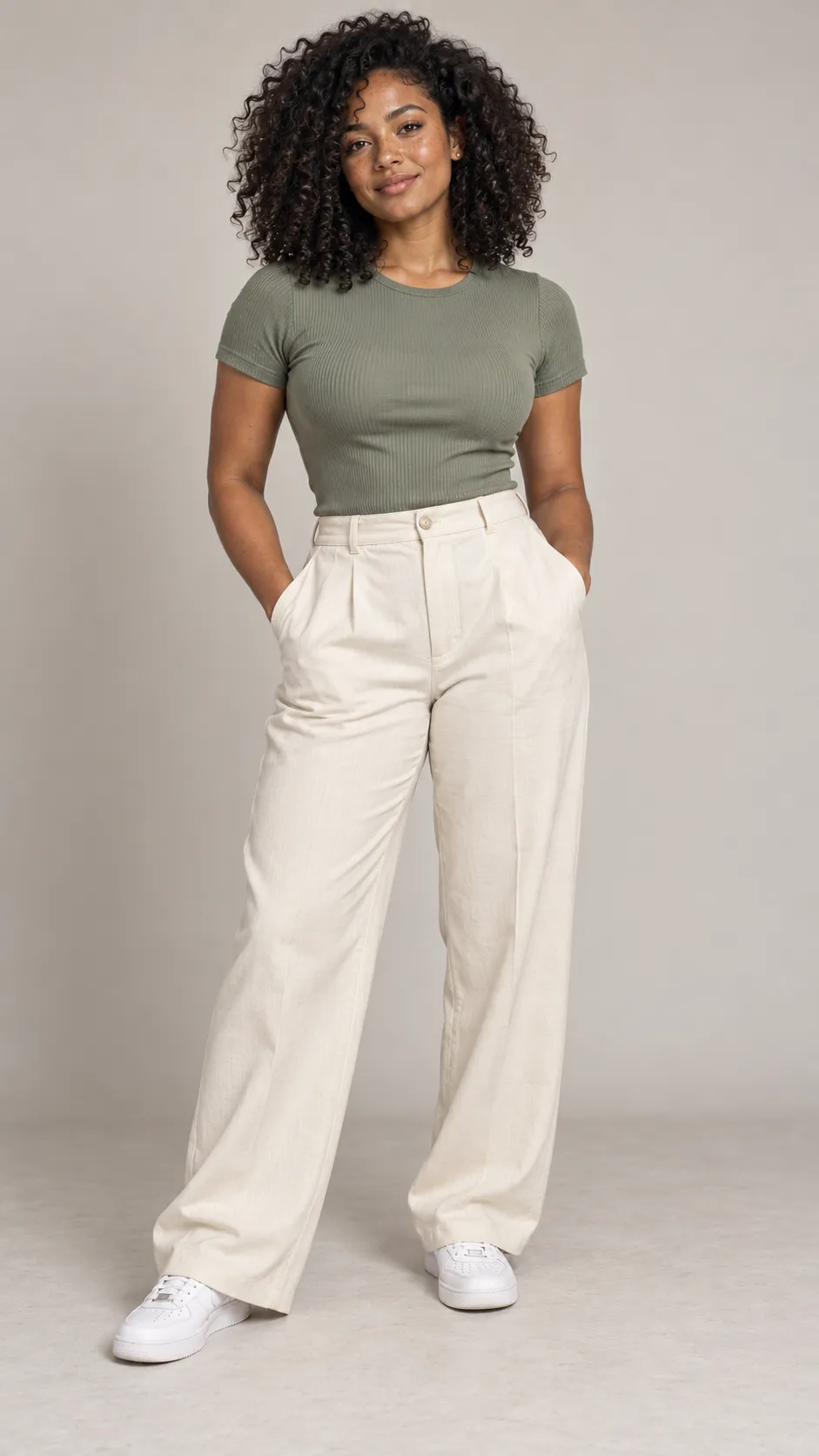 Generate a photorealistic full-body fashion portrait of a 26-year-old mixed-heritage woman, approximately 167 cm tall, with a medium athletic build, medium-deep brown skin, subtle freckles across her nose and cheeks, dark hazel eyes, and dense natural black curls cut into a rounded shoulder-length shape. She wears a muted-sage fitted top, high-waisted cream trousers, minimalist gold studs, and clean white sneakers. Her expression is confident and approachable, with natural makeup and realistic facial asymmetry. Preserve the complexity of her skin tone, individual curls, and natural body shape. Soft balanced studio lighting, pale warm-gray background, detailed fabric weave, gentle floor shadows, contemporary inclusive fashion catalog photography, full head-to-toe composition, both shoes visible, vertical 9:16. No text, no watermark, no skin lightening.